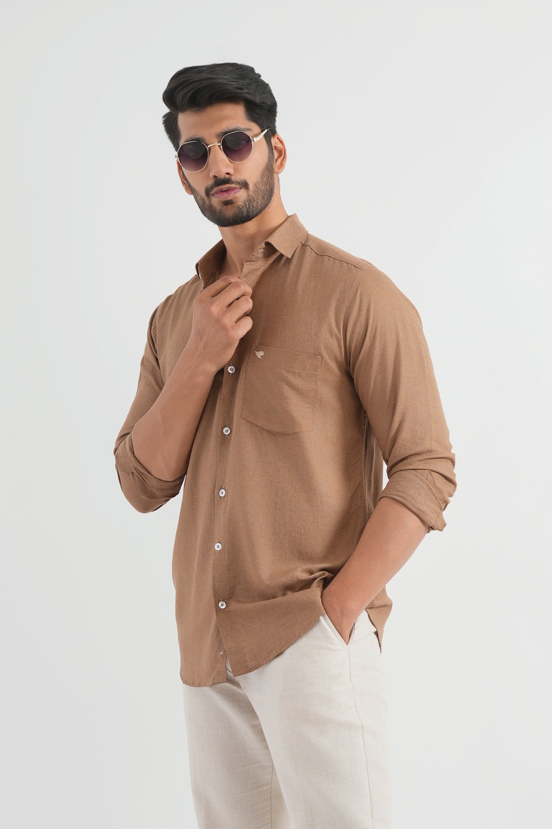 A man wearing a mocha brown linen shirt with white trousers and sunglasses, standing against a white background.