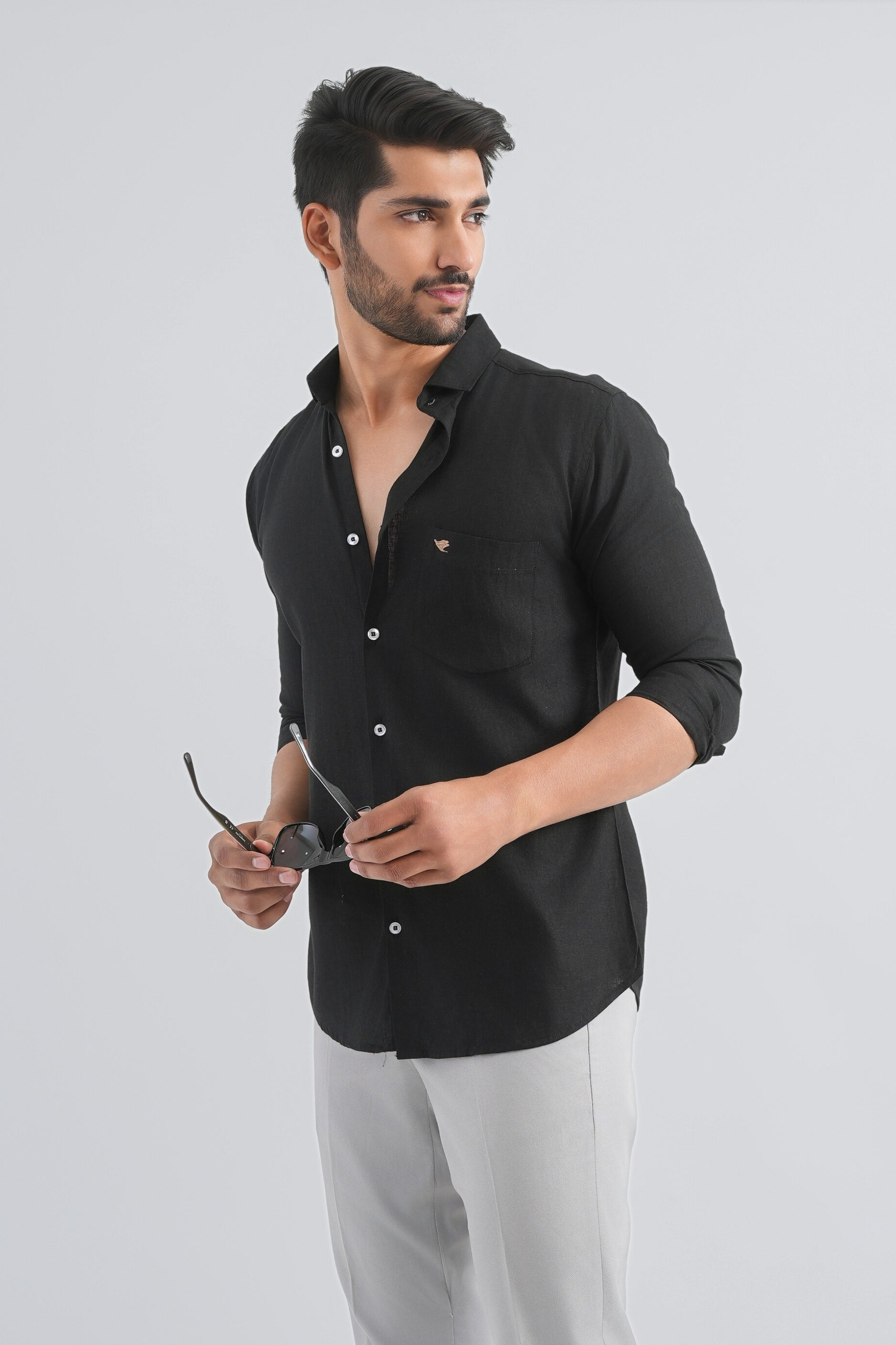 Man wearing a black shirt and light gray pants holding sunglasses on a white background