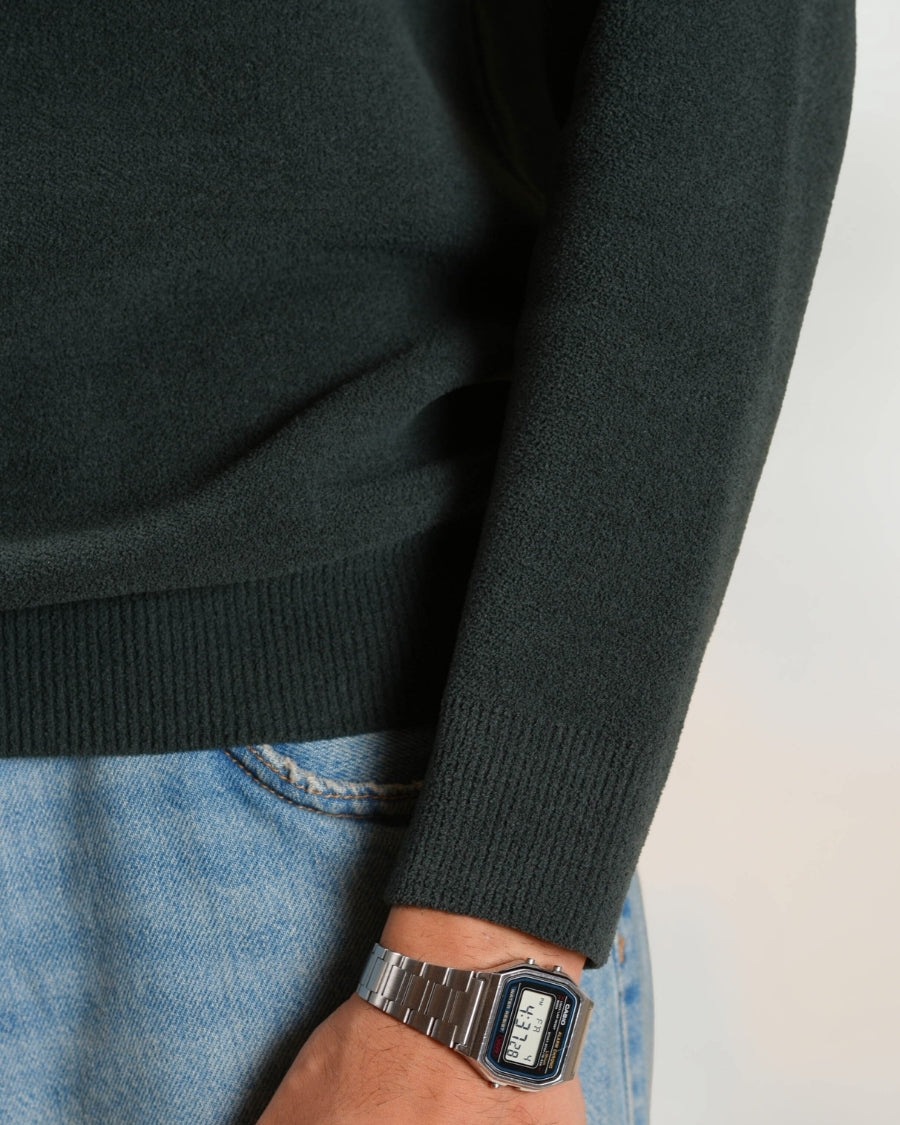Person wearing a dark green sweater and blue jeans with a silver watch on a white background