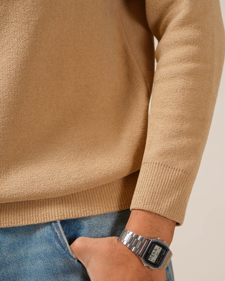 Person wearing a beige sweater and blue jeans with a silver watch on a plain background