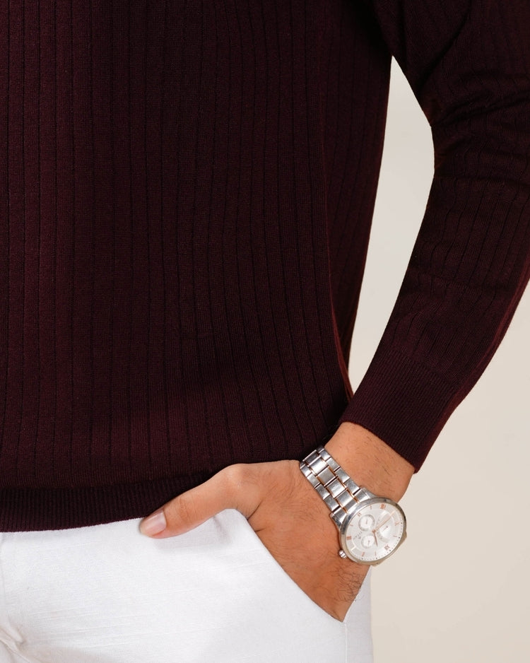 Person wearing a dark brown ribbed sweater and white pants with a silver watch on a neutral background