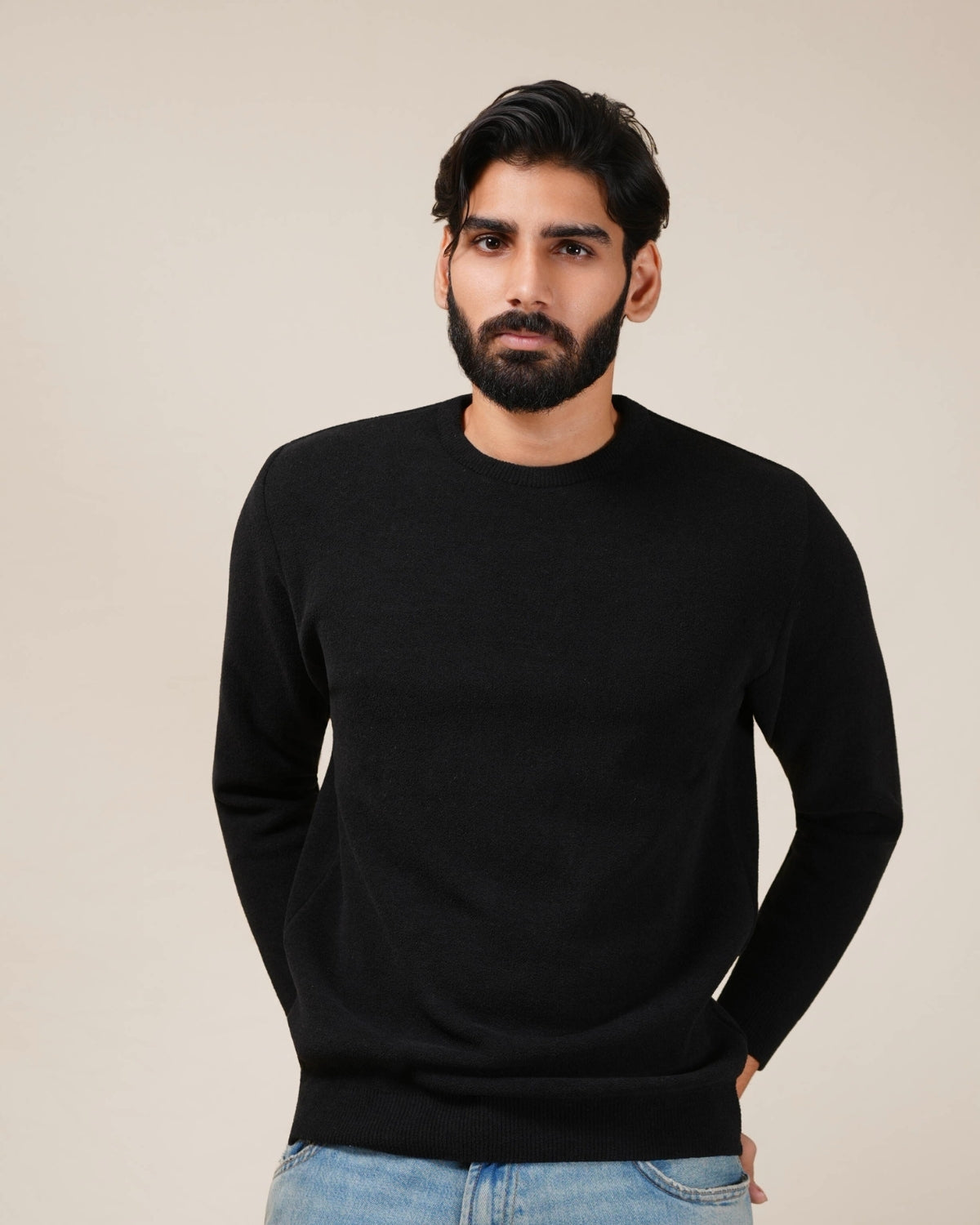 Man wearing a black sweater and light blue jeans against a beige background